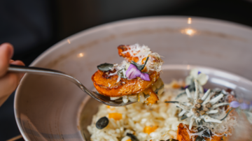 A plate with risotto and fried shrimp is visible. The dish is decorated with edible flowers and pumpkin seeds.