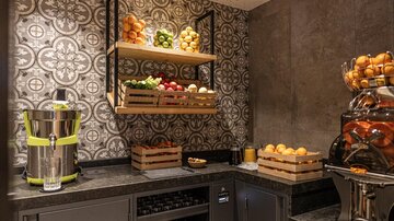 A stylish kitchen area with shelves full of fresh fruits and a beverage vending machine. The walls are decoratively designed with an appealing pattern.