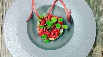 A artfully arranged dish on a gray plate. It contains fresh raspberries, vegetables, and decorative garnishes.