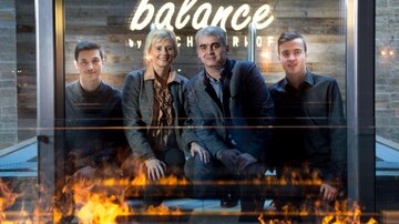 A group of four people is posing in a modern room in front of a glowing sign. In the foreground, there is a fire creating a cozy atmosphere.