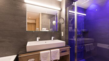 A modern bathroom with a large sink and stylish fixtures. The shower is separated by glass walls and is illuminated by blue light.