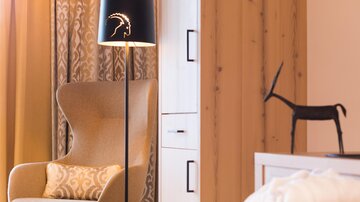 An elegant armchair stands beside a floor lamp in a modern room. In the background, wood paneling and a decorative object are visible.