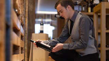 A young man is examining a wine bottle in a wine shop. He is wearing an elegant suit and kneeling in front of a shelf.