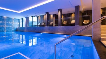 A luxurious indoor pool with clear blue water. The surroundings are modern and elegant with relaxing loungers.