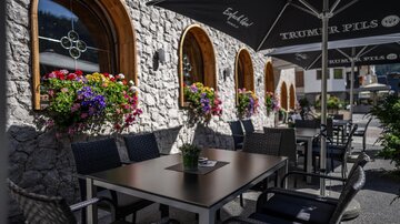 A cozy outdoor area with a table and chairs in front of a stone wall. Flowers in colorful boxes adorn the windows.