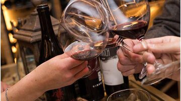 Several glasses of red wine toast with each other. In the background, wine bottles and corks are visible.