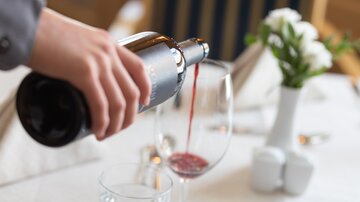 An elegant restaurant setting with a glass goblet that is currently being filled with wine. There is a vase with fresh flowers on the table.