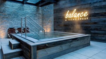 A modern wellness area with a jacuzzi and wooden paneling. The wall is designed with the glowing inscription "balance by Ischglerhof."