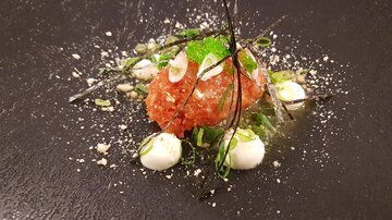 An artfully arranged dish with a main ingredient in the center, surrounded by green herbs and small white sauce dots. The presentation is on a black plate, which contrasts with the colors of the food.