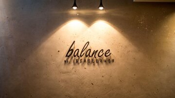A minimalist wall design featuring the inscription "balance" in elegant typography. Two ceiling lights illuminate the lettering.