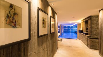 A modern designed hallway with wooden paneling and illuminated walls. At the end of the hallway, there is a view of an indoor pool with blue water.
