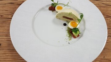 An elegant plate with a creative dish featuring a piece of terrine and two raw eggs. The dish is garnished with fresh herbs and a colorful sauce.