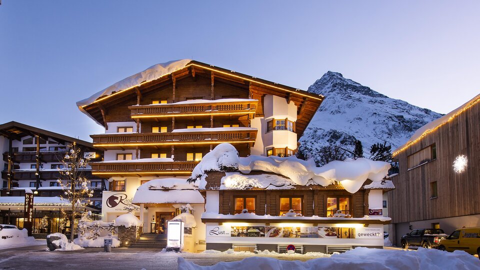 Ein charmantes Hotel im Schnee mit einem traditionellen Alpenstil. Im Hintergrund sind schneebedeckte Berge zu sehen.
