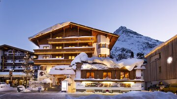 A charming hotel in the snow with a traditional Alpine style. Snow-covered mountains are visible in the background.