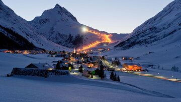 A snowy mountain landscape with a small village. The ski lifts and houses are gently illuminated in the twilight.