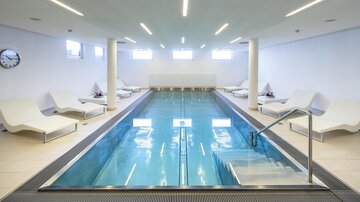 A modern swimming pool with clear water and loungers. The walls are bright and the lighting is friendly. | © Rössle