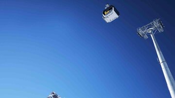 A cable car glides over snow-covered mountains under a clear blue sky. Nearby stands a tall light pole.