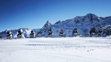 A snow landscape with several snow cannons against a clear blue sky. In the background, majestic mountains can be seen.