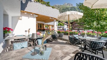 A cozy terrace with round tables and chairs. In the background, plants and mountains are visible, on a sunny day. | © Rössle