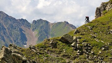 A mountain biker rides down a steep, rocky slope. The landscape features tall mountains and a green meadow in the background.