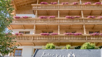 A beautiful hotel with wooden cladding and blooming balconies. The entrance is inviting and surrounded by plants.
