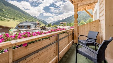 A cozy balcony with two chairs, surrounded by blooming flowers. In the background, there are green mountains and a blue sky with beautiful clouds.