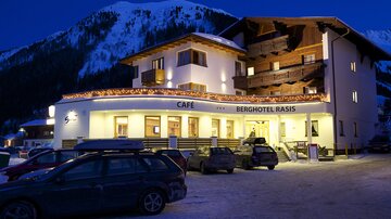 A cozy mountain hotel with a café, illuminated at dusk. Snow-covered mountains are visible in the background.