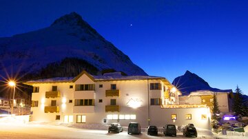 A cozy hotel in a snowy mountain landscape at night. The sky is dark blue and the mountains are majestically visible in the background.