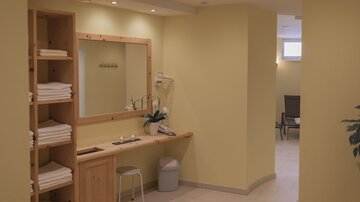 A cozy spa area with a wooden shelf full of towels. The room is decorated in warm yellow tones and features a mirror as well as plants.