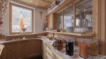 A cozy dining area with wooden paneling, a window view, and a buffet. Various drinks and fresh rolls are available on the counter.
