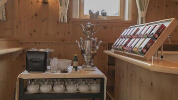 A table with a teapot and various types of tea in a wooden finish. In the background, some windows and a cozy atmosphere are visible.