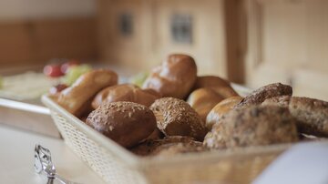 A basket with fresh rolls and pastries is on a wooden shelf. In the background, fresh ingredients and kitchen utensils can be seen.