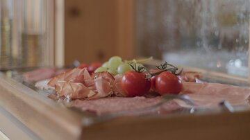 A serving tray filled with fresh ingredients including ham, tomatoes, and grapes. In the background, a wood-paneled wall can be seen.