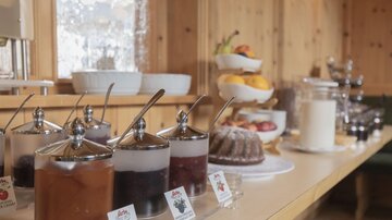 A buffet with various jams in glass containers and a fruit plate. In the background, additional snacks and drinks are visible.