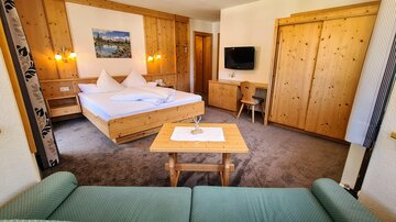 A cozy hotel room with wooden paneling and a large double bed. There is a small table, a television, and a couch.