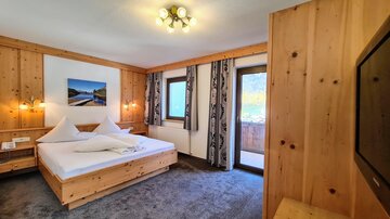 A cozy hotel room with wooden interiors and modern decorations. The room features a large bed, a television, and a balcony.