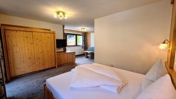 A cozy room with a double bed, wooden cabinets, and a television. The room has natural light and offers a view of the seating area.