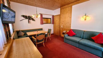 A cozy living space with a green sofa, a wooden table, and chairs. The walls are paneled with wood, and there is a television as well as charming lighting.