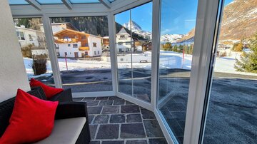 A bright conservatory with a view of snow-covered mountains and pretty houses. The room is furnished with a cozy seating area.