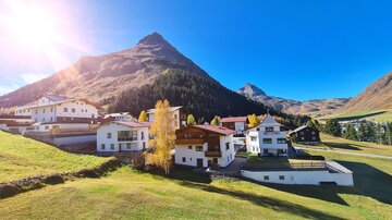 A picturesque mountain landscape with several buildings in a green valley. The sky is clear and sunny, with impressive mountains in the background.