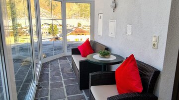 A bright conservatory with two black chairs and red cushions. The floor is designed with square stones, and large windows offer a view outside.