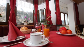 A cozy breakfast table with a red tablecloth, a cup of coffee, fresh rolls, a glass of orange juice, and fruit. In the background, there are flowers and large windows with a view of the mountains.