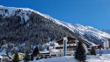 A wintry landscape with snow-covered mountains. In the foreground stands a charming hotel, surrounded by fir trees and a clear, blue sky.
