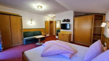 A cozy hotel room with a double bed, a sofa, and a table. The decor is made of wood and there is a television on the wall.