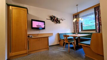 A cozy living space with a table and a corner bench. A television hangs on the wall and a window overlooks the outdoors.