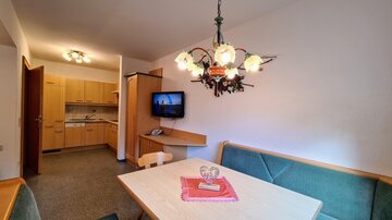A cozy dining area with a table and a green bench. The kitchen is visible in the background and there is a television on the wall.