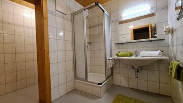A simple bathroom with a shower and a sink. The walls are tiled and there is a small window for daylight.