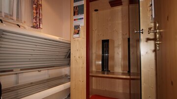 An interior with a sunbed and a sauna. The walls are made of wood and there is a glass door to the sauna.