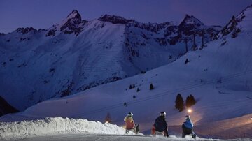 Three skiers are sitting on a snowy slope, looking at the mountains. The sky is dark blue, and the snowy landscape has a peaceful, wintry atmosphere.