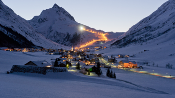 A picturesque winter landscape with a small village surrounded by snow-covered mountains. The lights of the village twinkle in the twilight as the ski slope is gently illuminated.